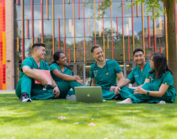 Five medical students wearing green scrubs sit on a campus lawn talking and studying.