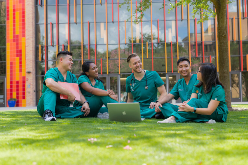 Five medical students wearing green scrubs sit on a campus lawn talking and studying.
