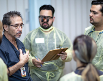 A faculty member in blue scrubs instructs a group of dental students who are gowned up and ready for patient care.