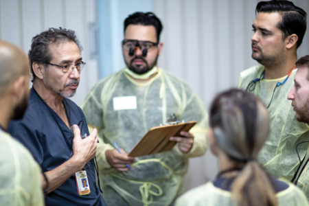 A faculty member in blue scrubs instructs a group of dental students who are gowned up and ready for patient care.
