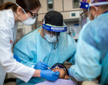 A dental faculty member leans over the shoulder of a dental resident to offer guidance during a procedure.