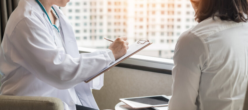 A stock photo of a doctor in a white coat with a clipboard speaking with a patient.