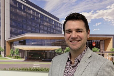Tyler Witges, MHA, MLS(ASCP), hospital laboratory operations director for the UT Health San Antonio Multispecialty and Research Hospital, stands in front of a rendering of the hospital.