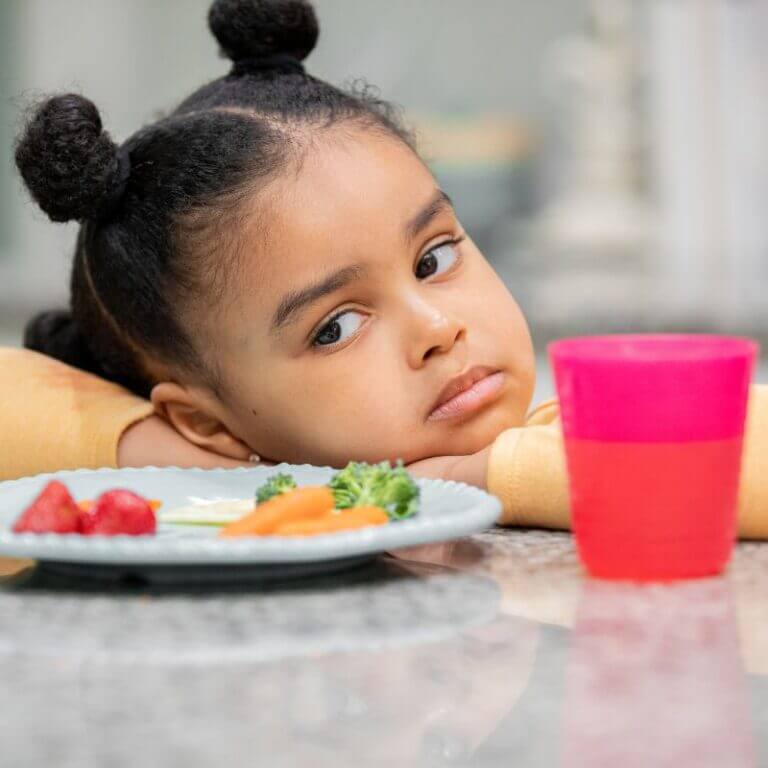 A toddler resting her head on the table refusing to eat her food.