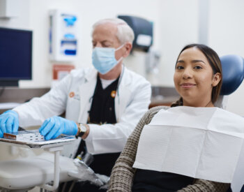 A patient sitting in a dental chair smiles while the dentist behind her is arranging dental tools for an exam.