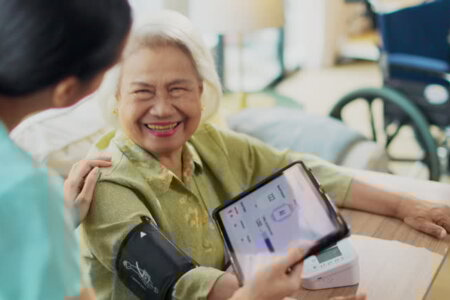 Woman asian nurse taking blood pressure of a senior woman patient at home.Home care healthcare professional using digital tablet