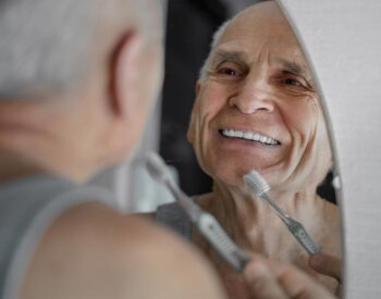 An older male smiles into the mirror while bringing a toothbrush up to his mouth.