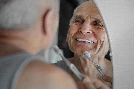 An older male smiles into the mirror while bringing a toothbrush up to his mouth.