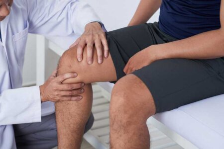 A physician in a white coat examines a patient wearing black shorts by squeezing his knee.