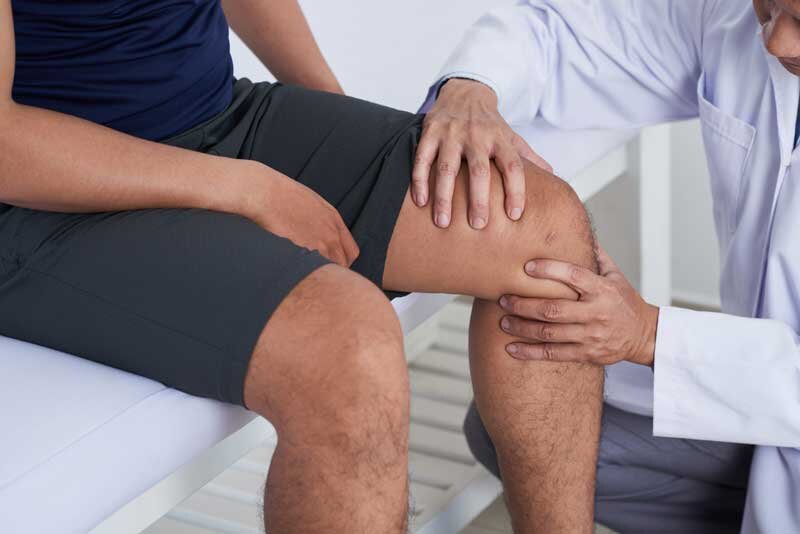 A physician in a white coat examines a patient wearing black shorts by squeezing his knee.