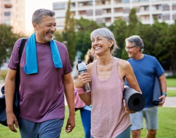 A woman carrying a yoga mat and water bottle talks with a man carrying a gym bag and wearing a towel around his neck.