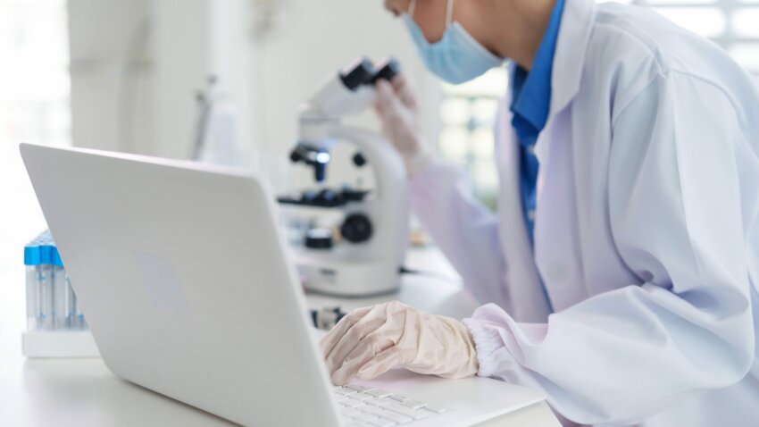Scientist conducting medical research in a lab. Expert working on a laptop while observing through a microscope. Focus professional dedication.