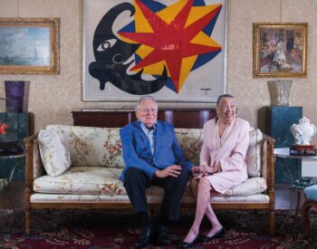 Joe R. and Teresa Lozano Long sit on a couch in front of a colorful painting.
