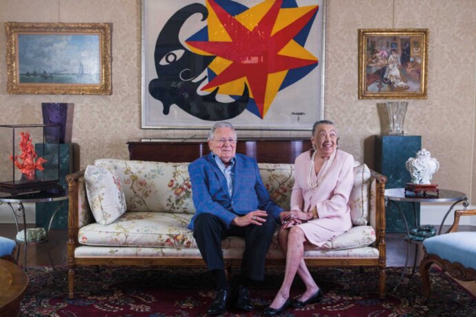 Joe R. and Teresa Lozano Long sit on a couch in front of a colorful painting.